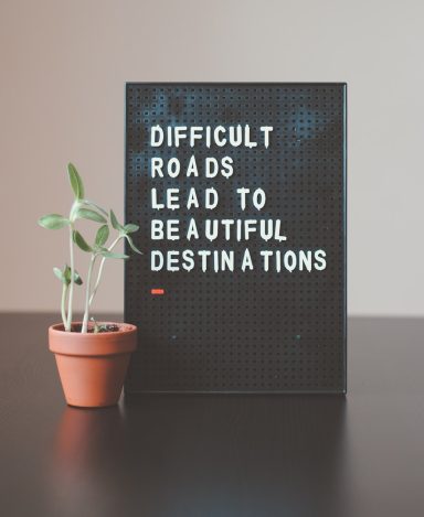 Motivation Ein schwarzes Schild mit dem Text „Difficult roads lead to beautiful destinations“ und einem Topf mit Pflanze.